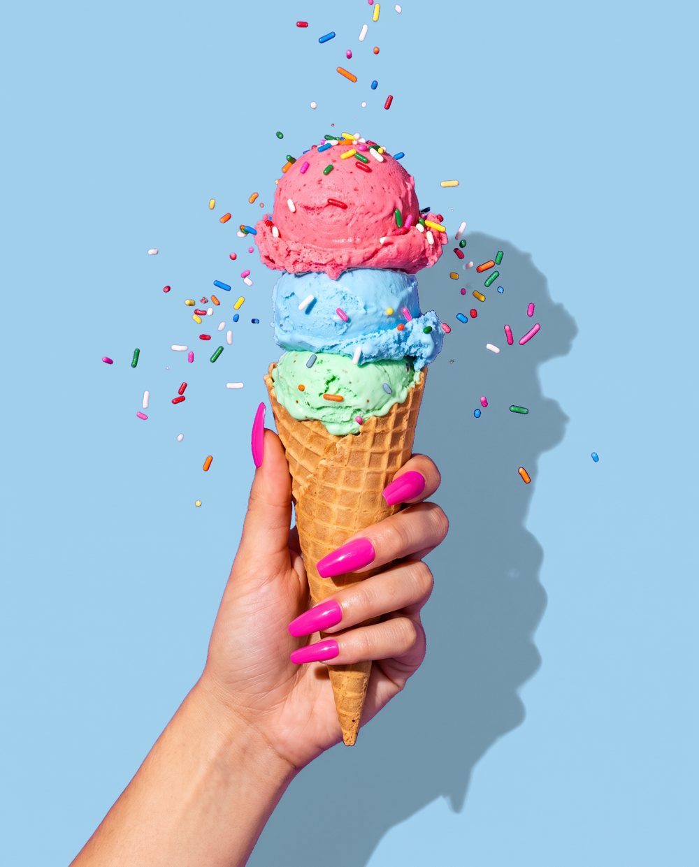 Hand holding a triple scoop cone with sprinkles flying on a violet background
